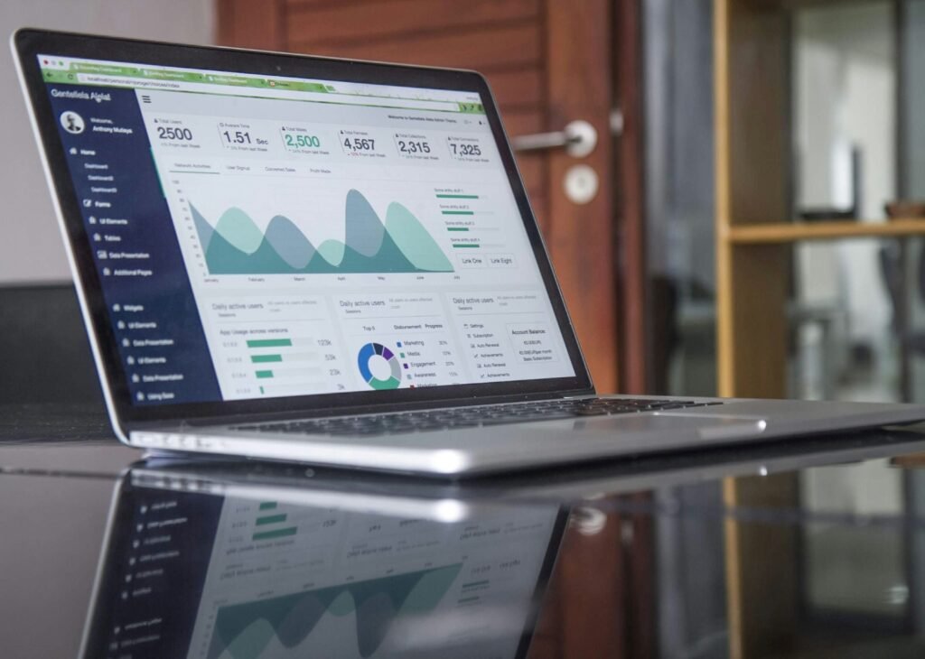A laptop displaying an analytics dashboard with graphs, statistics, and data visualizations, placed on a reflective glass table in an office setting. A wooden door and a bookshelf are visible in the background.